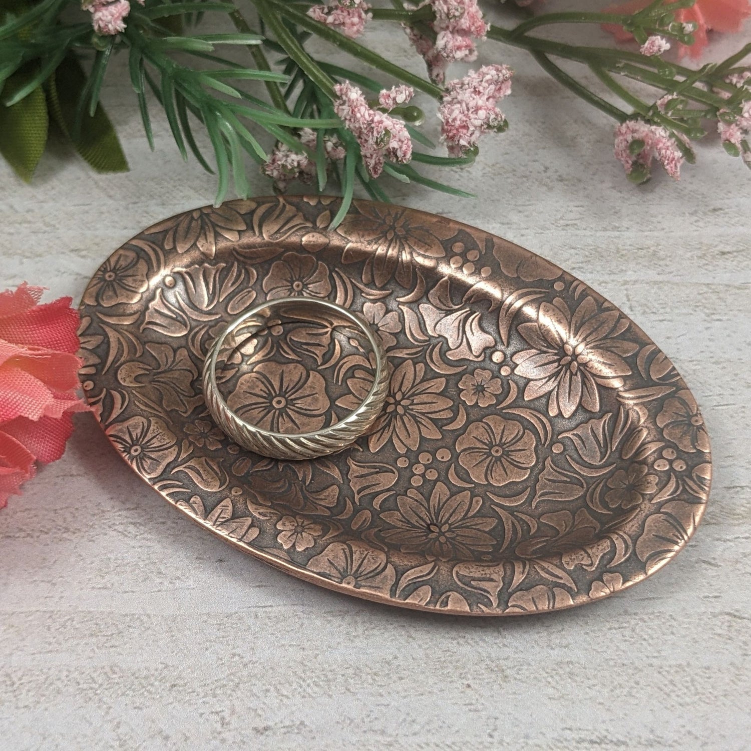 Oval copper ring dish with raised edge. Design of a variety of garden flowers, leaves, and berries covers the entire surface. Staged with a ring and flowers in the background.