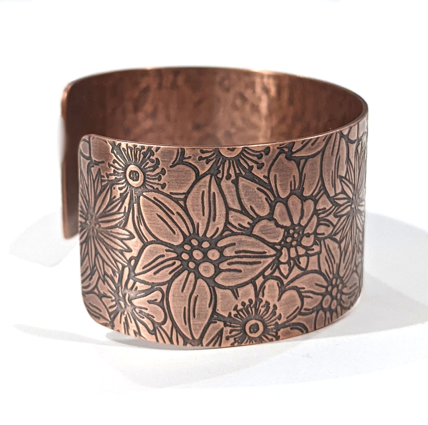 Wide copper bracelet with impressed design of flowers. The entire cuff is covered in various kinds of flowers in full bloom. Side view