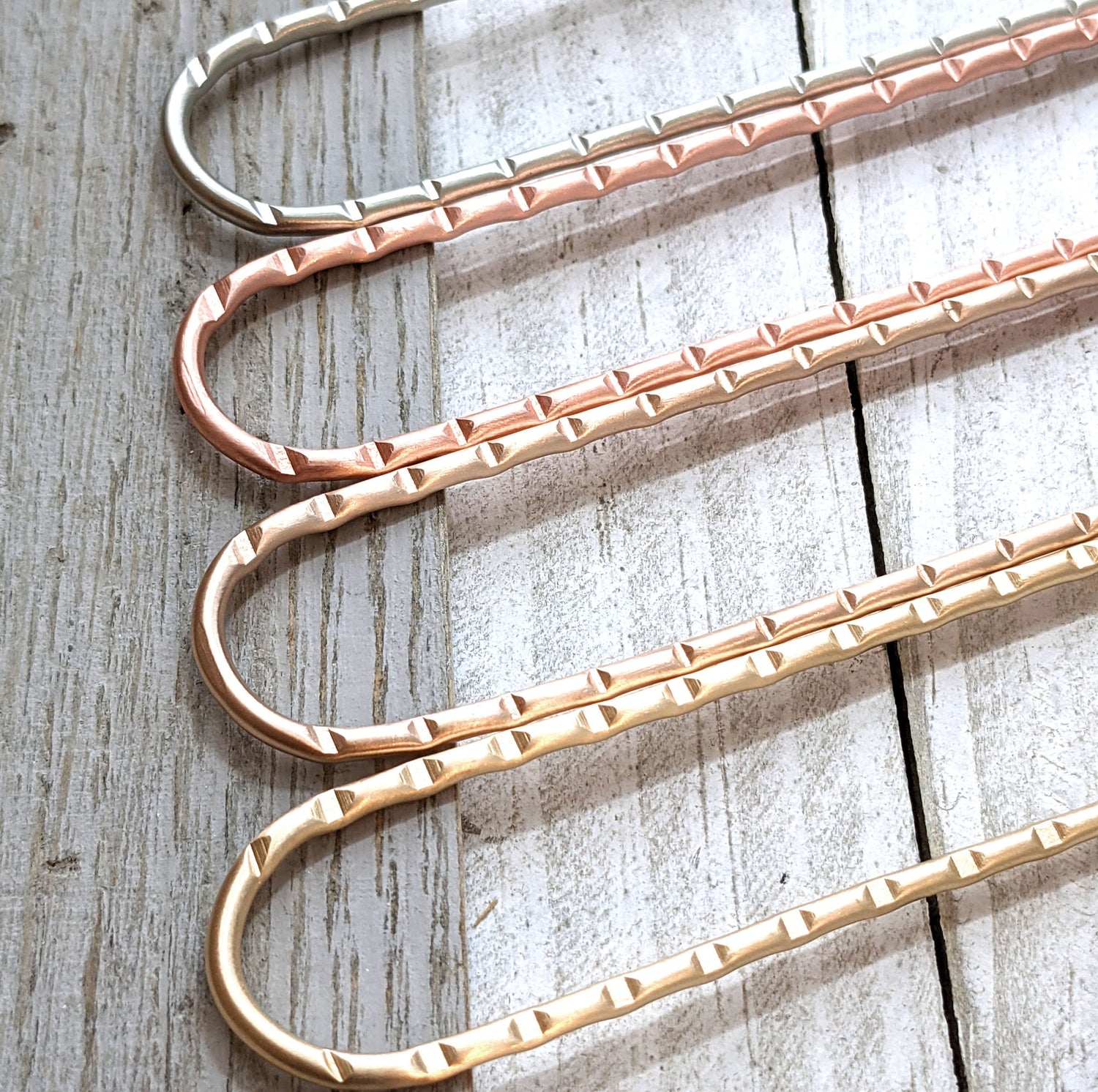 Metal hair forks shown in the four options - nickel, copper, bronze, and brass. This picture shows the detailed deep line grooves in the top portion of each hair stick.