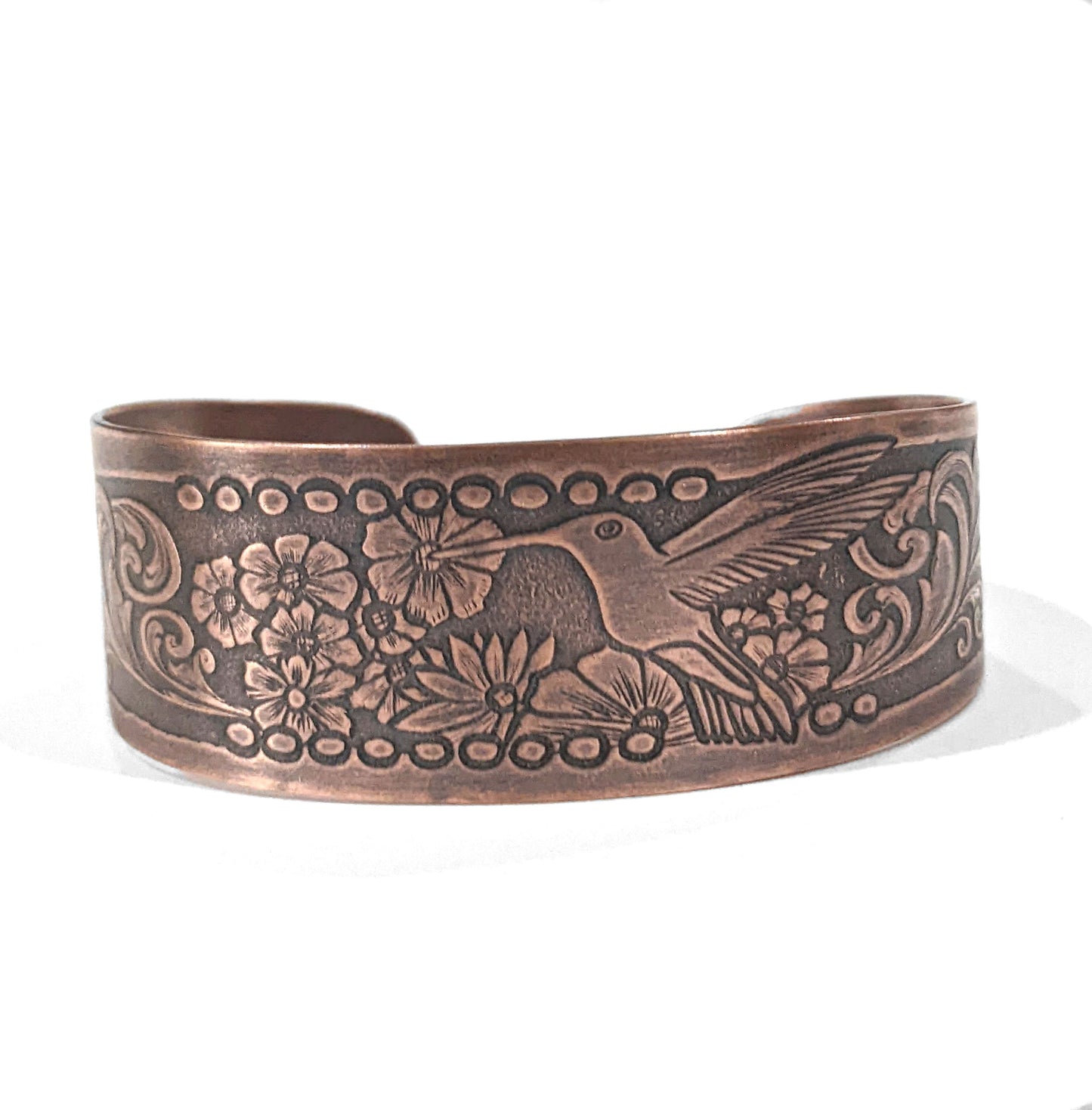 Copper cuff bracelet with hummingbirds and flowers. The main scene is a hummingbird in flight drinking nectar from one of a field of flowers. At each end of the cuff there is a smaller hummingbird drinking from a single flower. The cuff is tapered, just under and inch wide at the center tapering to one half inch at the ends.