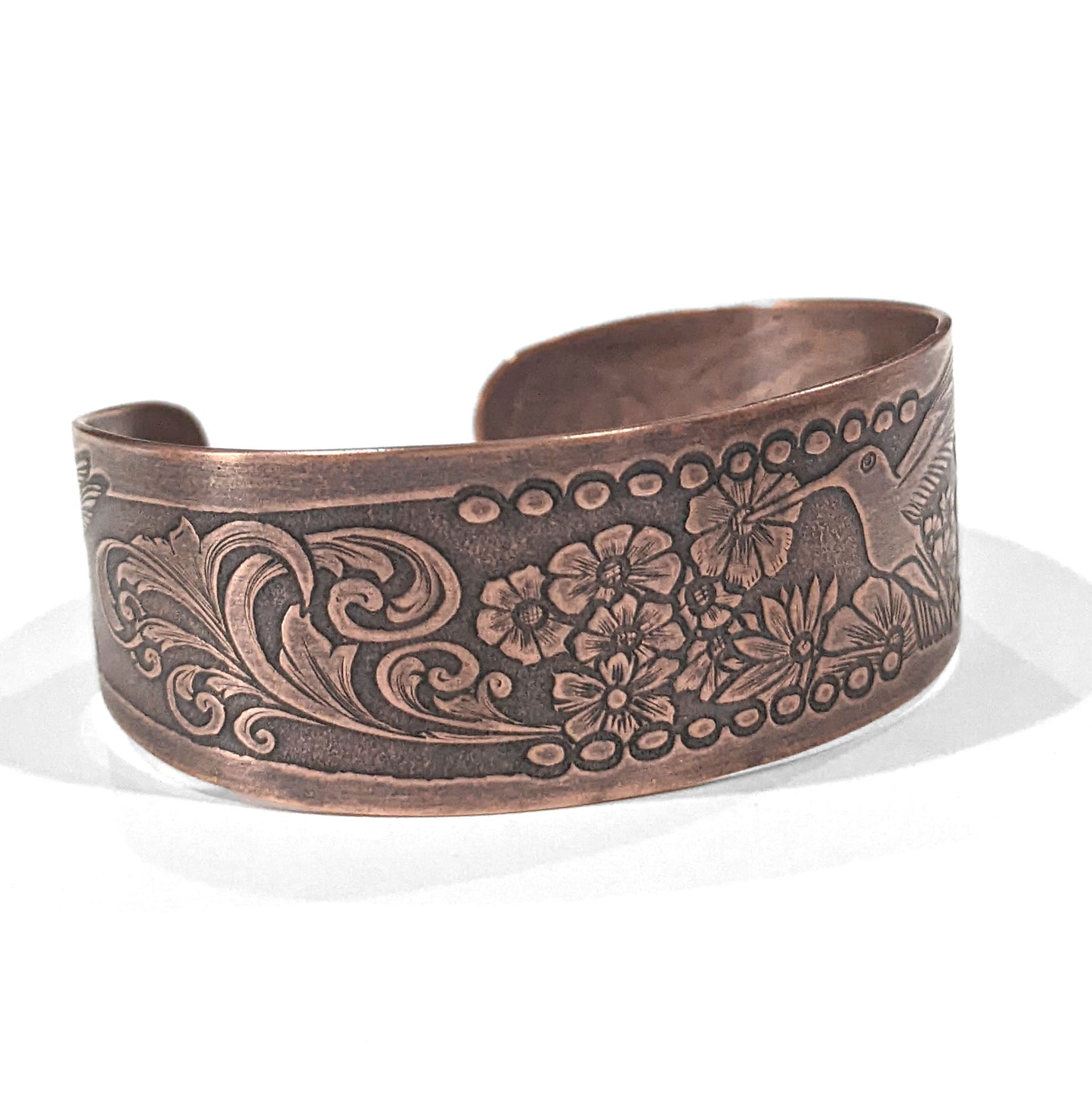 Copper cuff bracelet with hummingbirds and flowers. The main scene is a hummingbird in flight drinking nectar from one of a field of flowers. At each end of the cuff there is a smaller hummingbird drinking from a single flower. The cuff is tapered, just under and inch wide at the center tapering to one half inch at the ends.