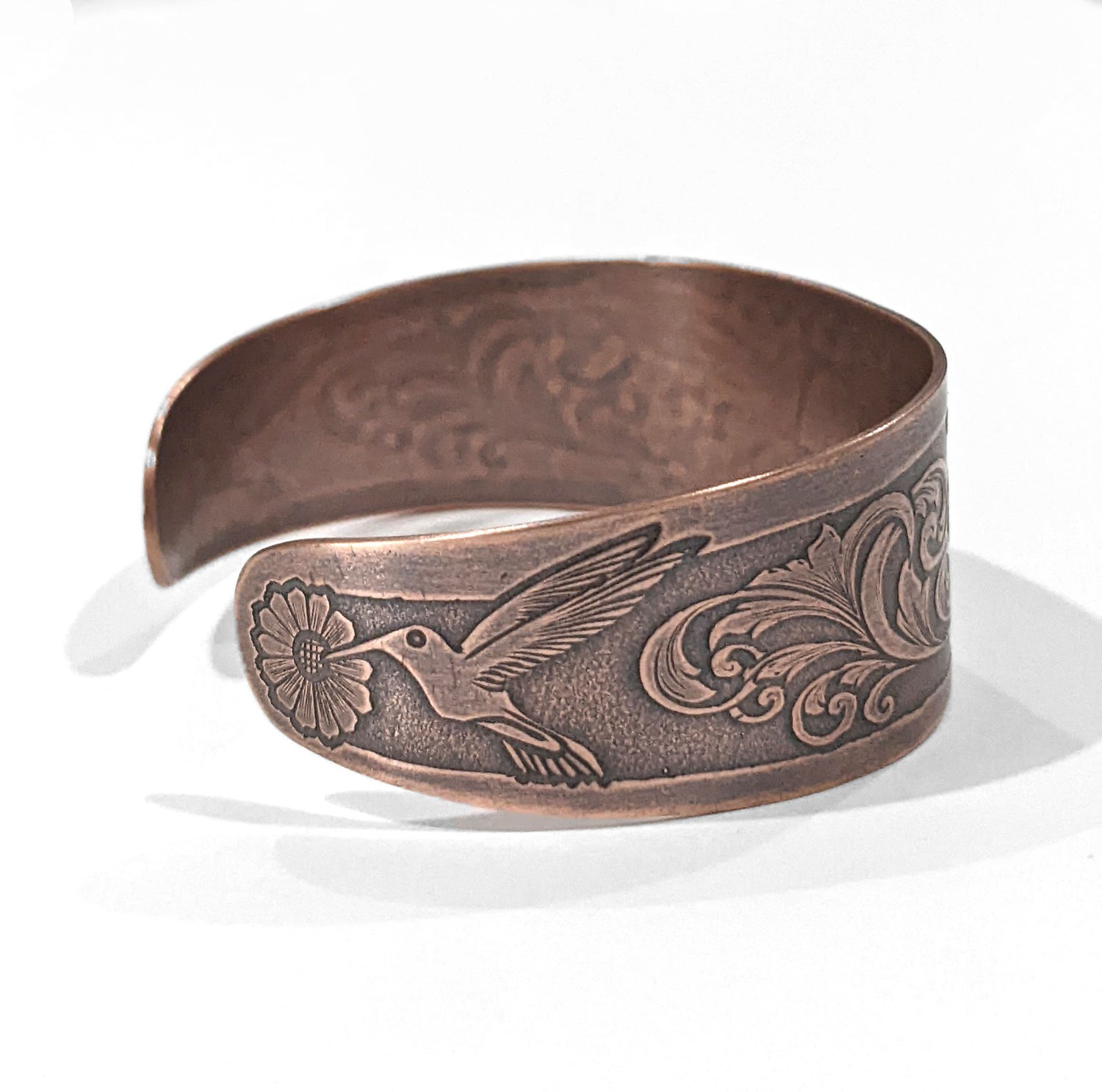 Copper cuff bracelet with hummingbirds and flowers. The main scene is a hummingbird in flight drinking nectar from one of a field of flowers. At each end of the cuff there is a smaller hummingbird drinking from a single flower. The cuff is tapered, just under and inch wide at the center tapering to one half inch at the ends. This is side view showing the smaller hummingbird.