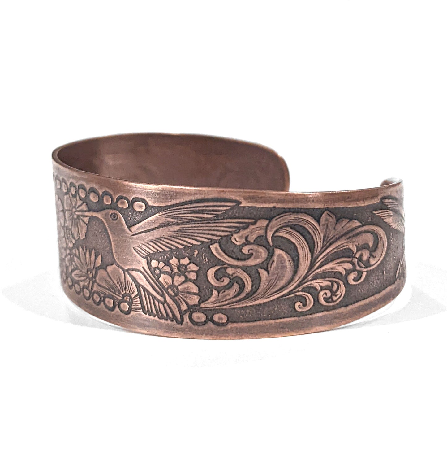 Copper cuff bracelet with hummingbirds and flowers. The main scene is a hummingbird in flight drinking nectar from one of a field of flowers. At each end of the cuff there is a smaller hummingbird drinking from a single flower. The cuff is tapered, just under and inch wide at the center tapering to one half inch at the ends. This is side view showing a flourish design.