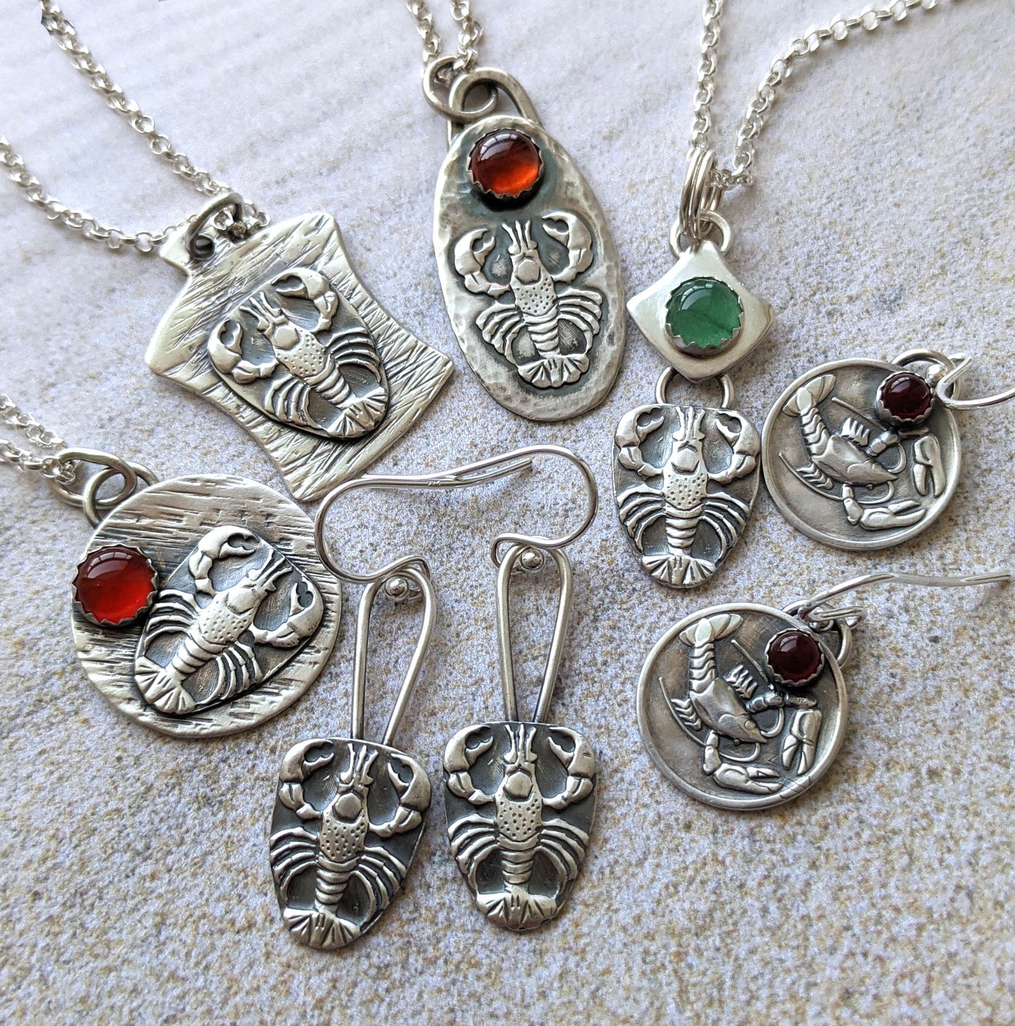 Collection of lobster jewelry. Showing several sterling silver pendants and earrings. There are two different lobster designs and some of the pendants and earrings have gemstone cabochons.