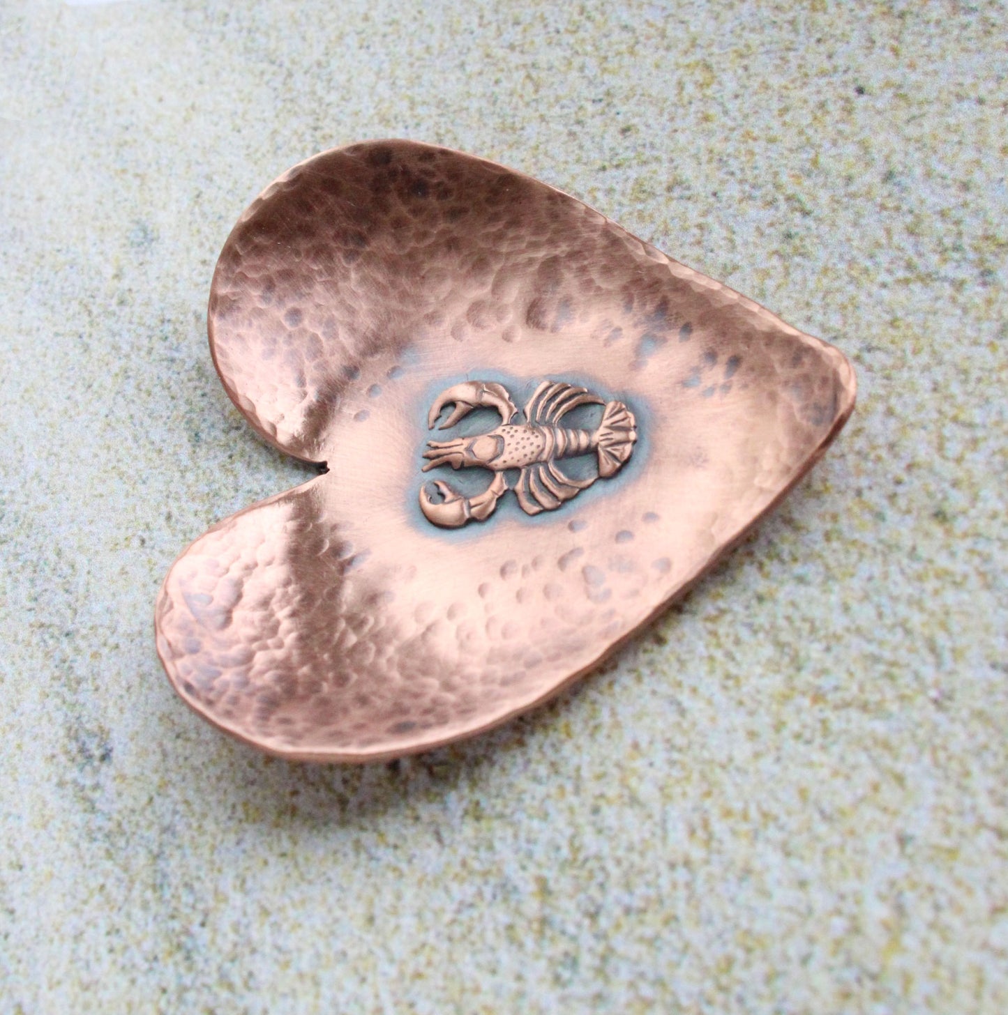 A small 2 inch heart shaped copper ring or trinket dish. There is a raised detailed impression of a lobster in the middle and hammered texture around the edge. The bottom is flat and the three points of the heart are gently curved up. The bowl is oxidized to enhance the details of the lobster and the hammer marks.