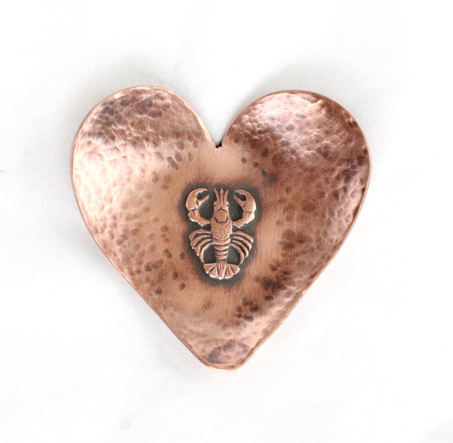 A small 2 inch heart shaped copper ring or trinket dish. There is a raised detailed impression of a lobster in the middle and hammered texture around the edge. The bottom is flat and the three points of the heart are gently curved up. The bowl is oxidized to enhance the details of the lobster and the hammer marks.