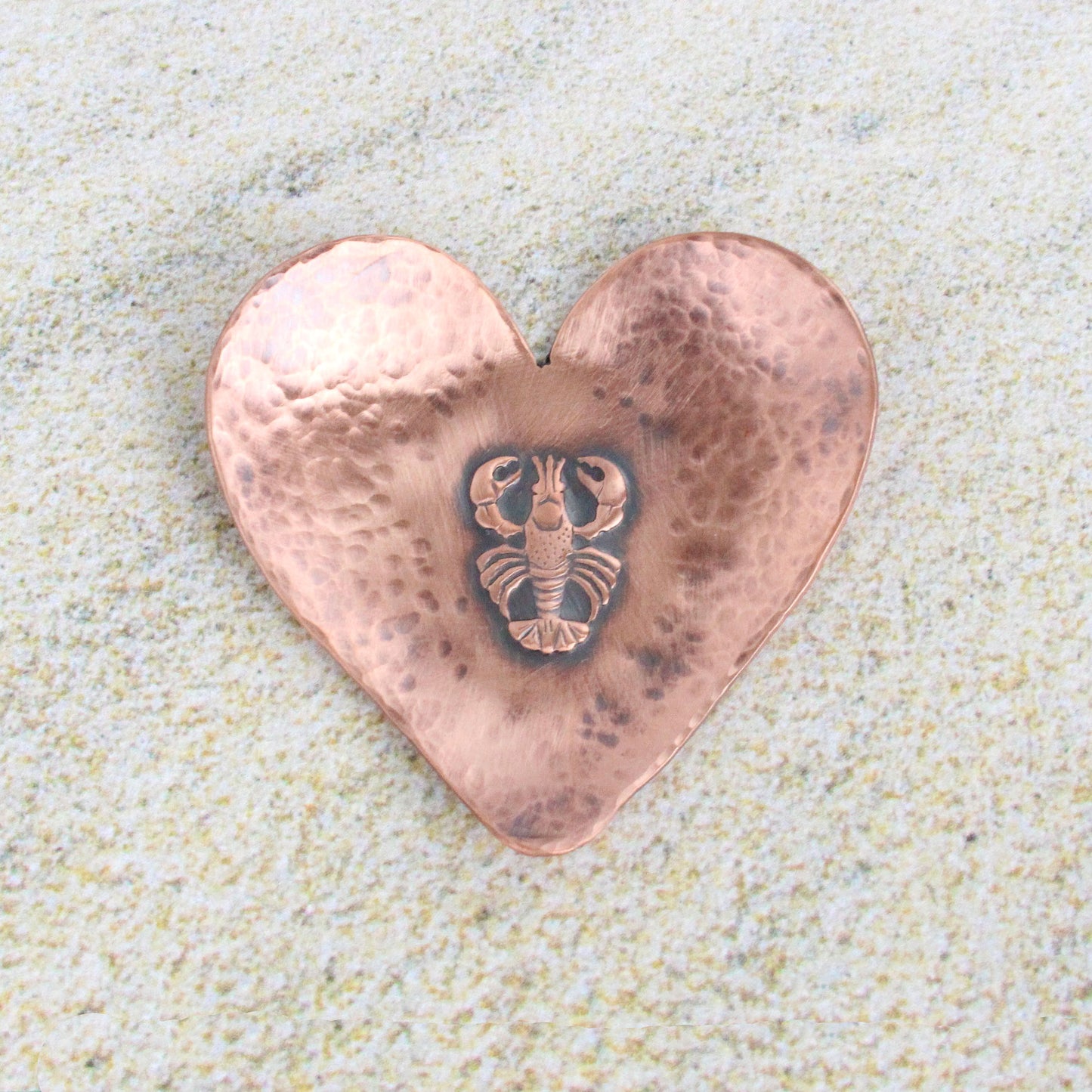 A small 2 inch heart shaped copper ring or trinket dish. There is a raised detailed impression of a lobster in the middle and hammered texture around the edge. The bottom is flat and the three points of the heart are gently curved up. The bowl is oxidized to enhance the details of the lobster and the hammer marks.