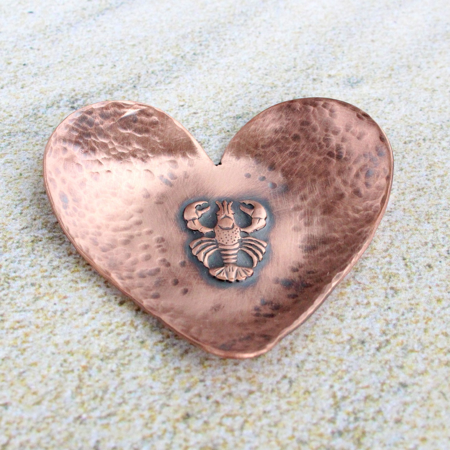 A small 2 inch heart shaped copper ring or trinket dish. There is a raised detailed impression of a lobster in the middle and hammered texture around the edge. The bottom is flat and the three points of the heart are gently curved up. The bowl is oxidized to enhance the details of the lobster and the hammer marks.