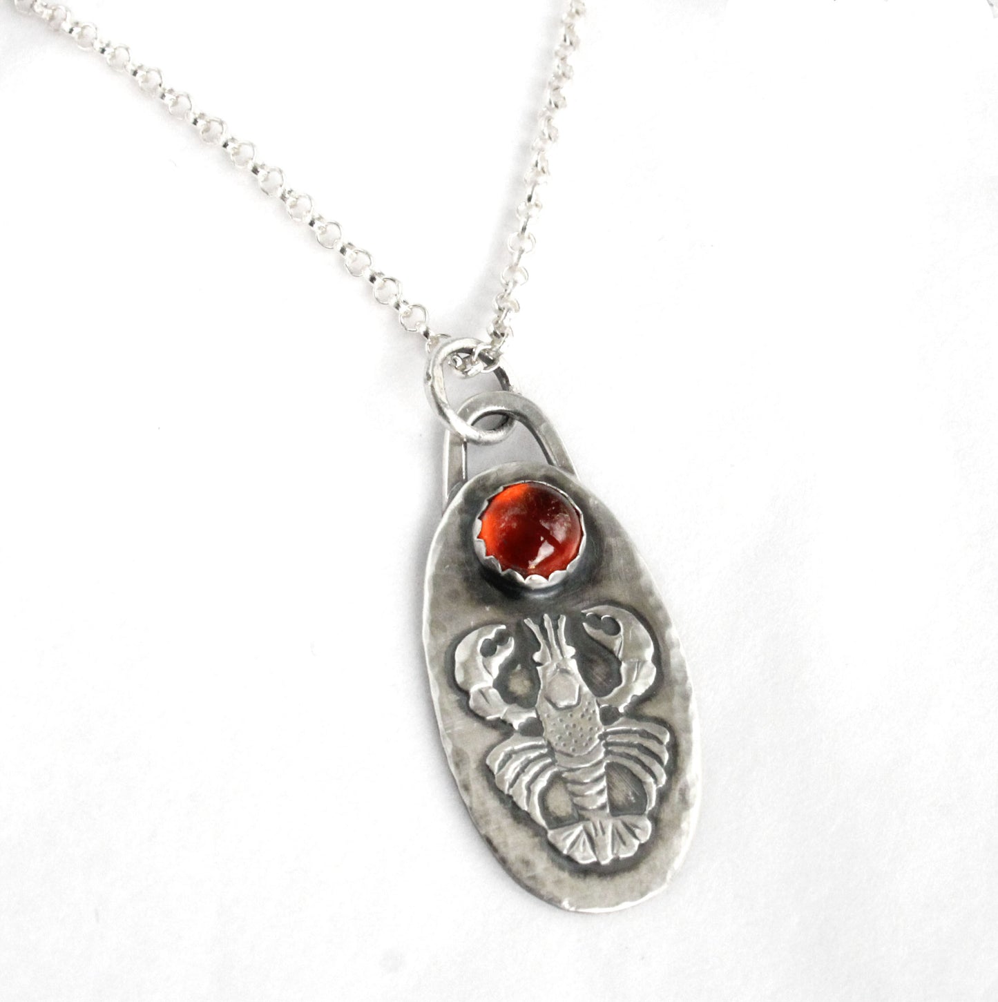 Lobster Sterling Silver Pendant with Hessonite Garnet. The pendand has a detailed raised impression of a lobster. Above the lobster is a 6mm hessonite garnet stone. The pendant is 1-1/4 inches long and about 5/8 inches wide and comes on an 18 inch sterling silver rolo chain.