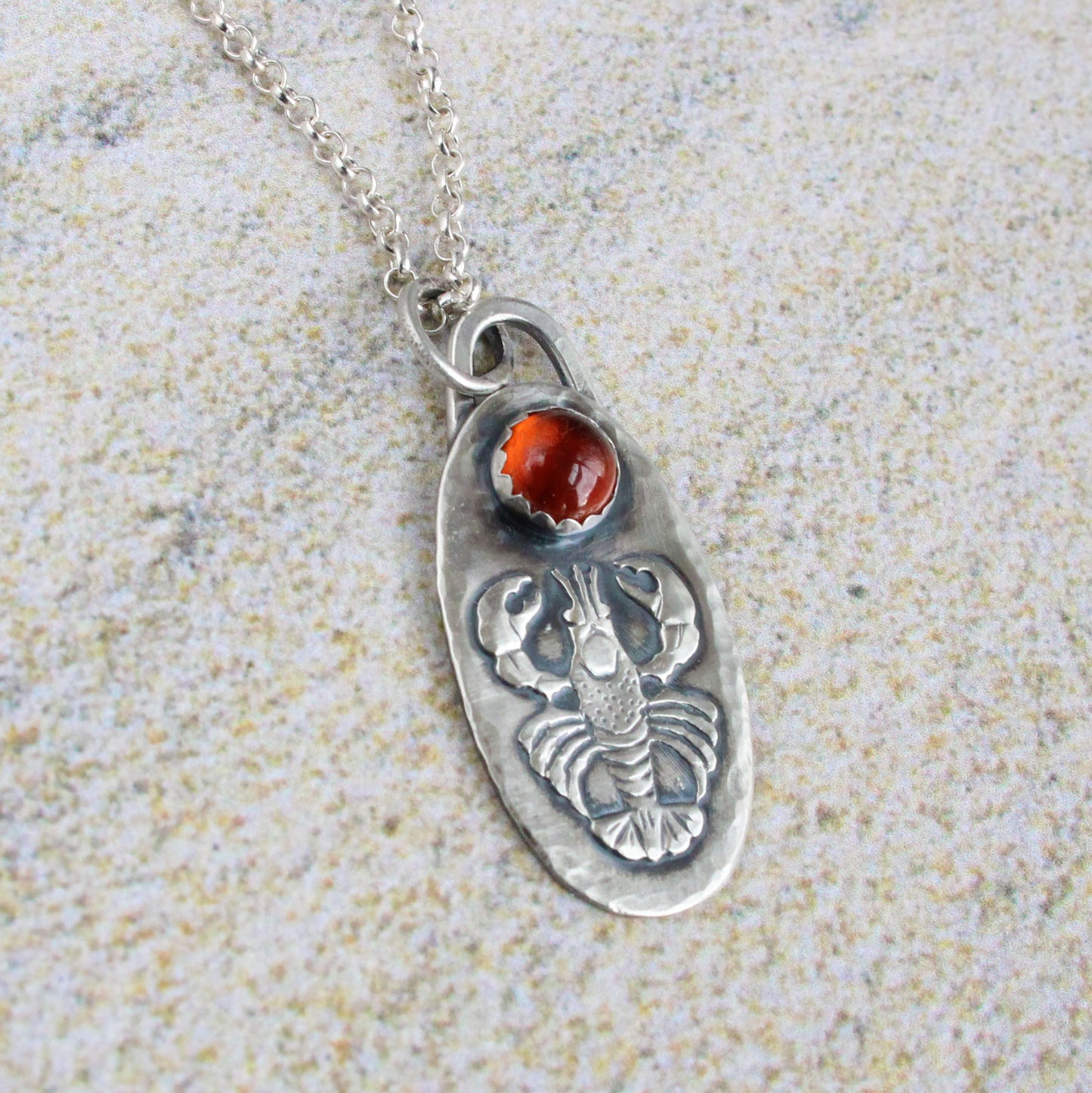 Lobster Sterling Silver Pendant with Hessonite Garnet. The pendand has a detailed raised impression of a lobster. Above the lobster is a 6mm hessonite garnet stone. The pendant is 1-1/4 inches long and about 5/8 inches wide and comes on an 18 inch sterling silver rolo chain.