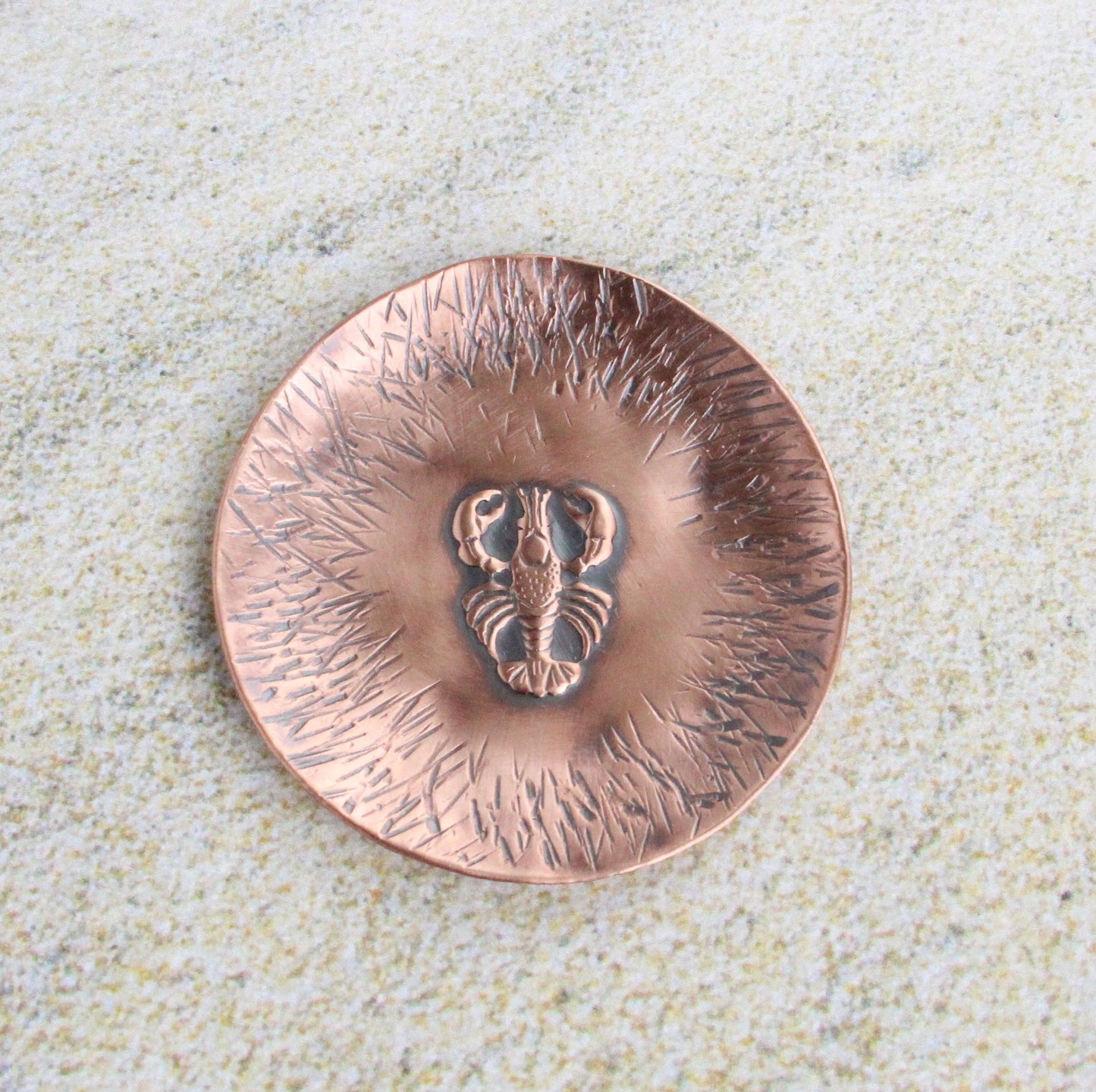 A 1-3/4 inch round copper ring or trinket dish. There is a raised detailed impression of a lobster in the middle and hammered lines around the edge. The bottom is flat and the sides are gently sloped. The bowl is oxidized to enhance the details of the lobster and the lines.