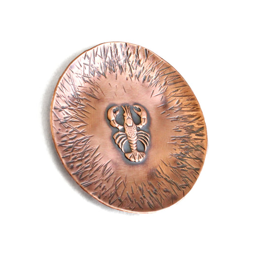 A small 1-3/4 inch round copper ring or trinket dish. There is a raised detailed impression of a lobster in the middle and hammered lines around the edge. The bottom is flat and the sides are gently sloped. The bowl is oxidized to enhance the details of the lobster and the lines.