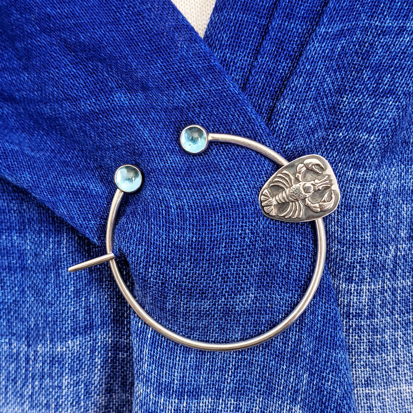 Sterling silver penannular brooch. The tip of the stem is decorated with a detailed dimensional lobster, and the ends of the loop have bright swiss blue topaz gemstone cabochons. Shown modeled on a blue shawl.