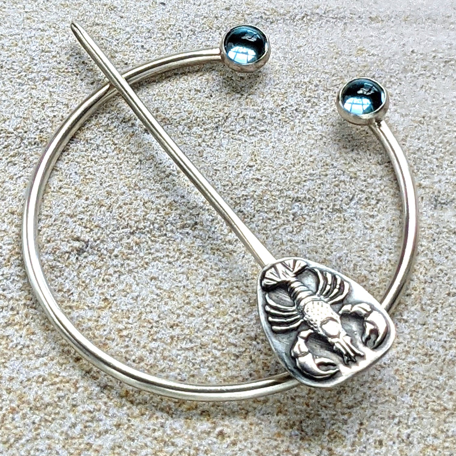 Sterling silver penannular brooch. The tip of the stem is decorated with a detailed dimensional lobster, and the ends of the loop have bright swiss blue topaz gemstone cabochons.