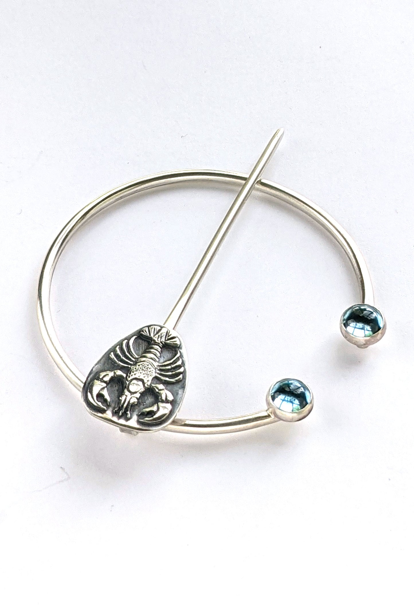 Sterling silver penannular brooch. The tip of the stem is decorated with a detailed dimensional lobster, and the ends of the loop have bright swiss blue topaz gemstone cabochons.