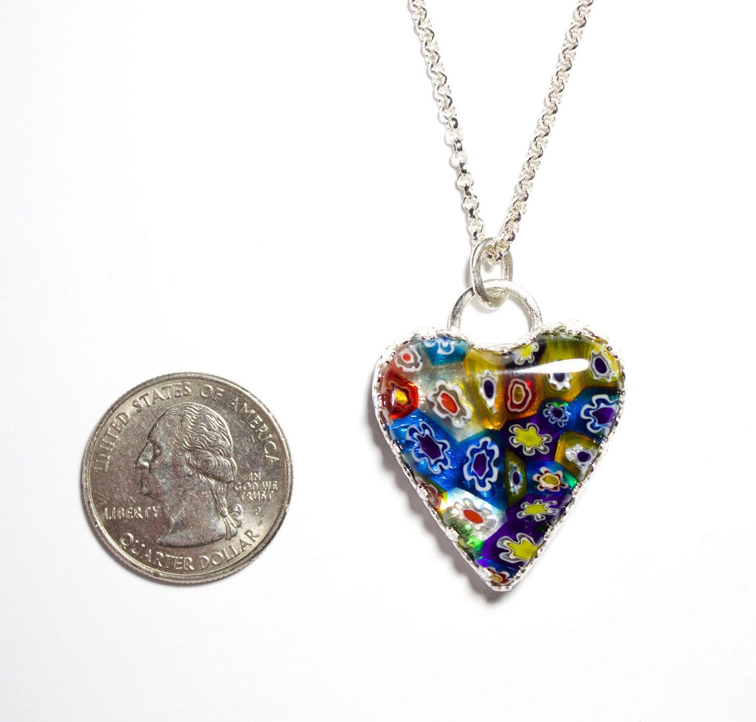 Heart shaped pendant made of millifiore glass. The heart is clear glass filled with glass tubes that are shaped like flowers and go from the front to the back of the heart. Multi color includes red, blue, yellow, and white. Surrounded by a heavy detailed gallery wire.