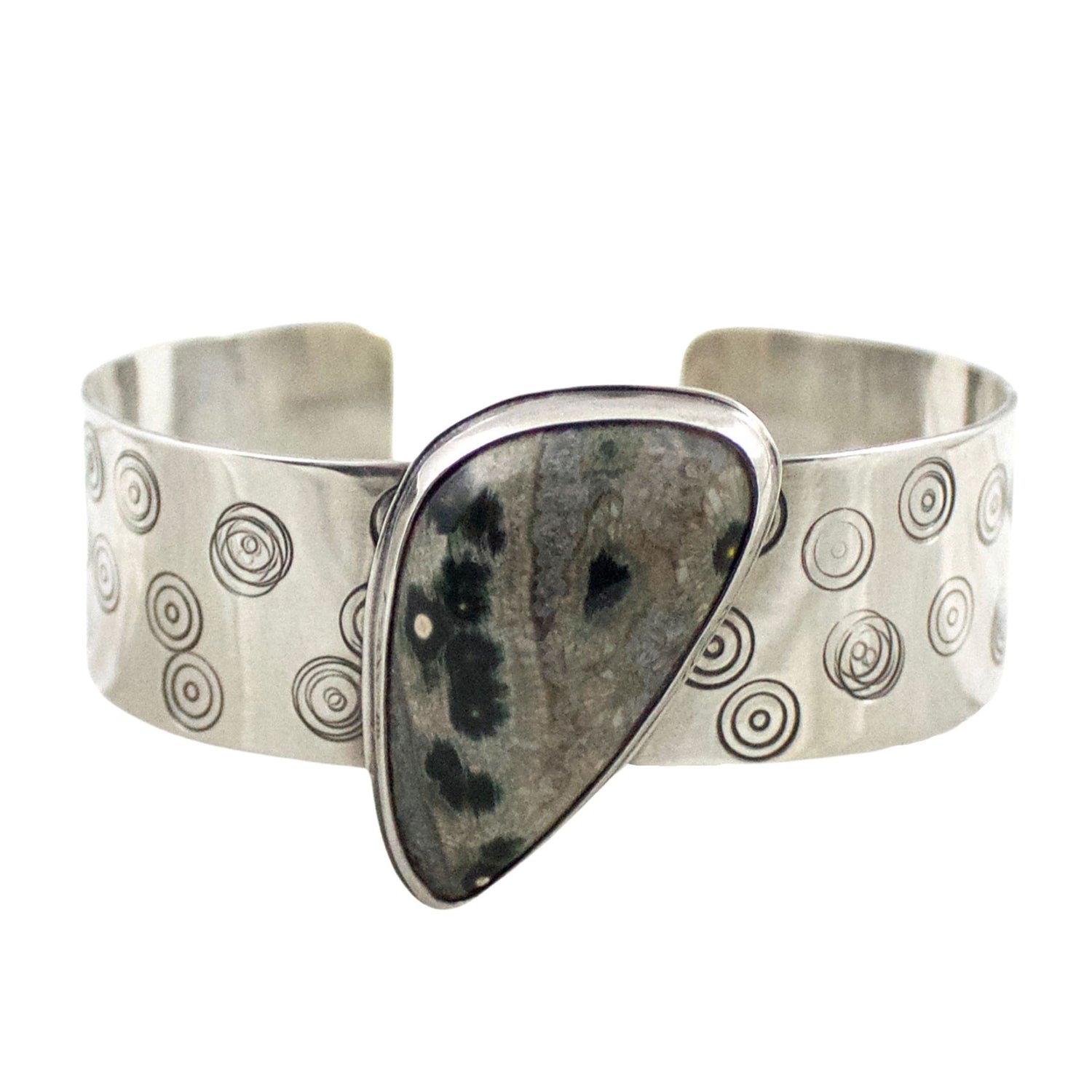 Sterling silver cuff bracelet with ocean jasper and orbs design. The cuff has an offset triangle shaped ocena jasper stone. The stone is muted browns and grays with round green circles throughout the stone. The circles are a feature of ocean jasper. The wide cuff bracele is stamped with nested circle patterns.