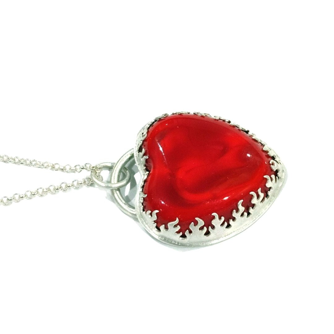 Pendant made from a large and thick lampwork glass stone in the shape of a heart. The heart is bright and bold red with swirls of lighter and darker reds, and is very shiny. The bezel that holds the glass stone is a cutout pattern of flames. All of the metal is sterling silver, and the pendant comes on a sterling silver necklace. Pictured from a side view.