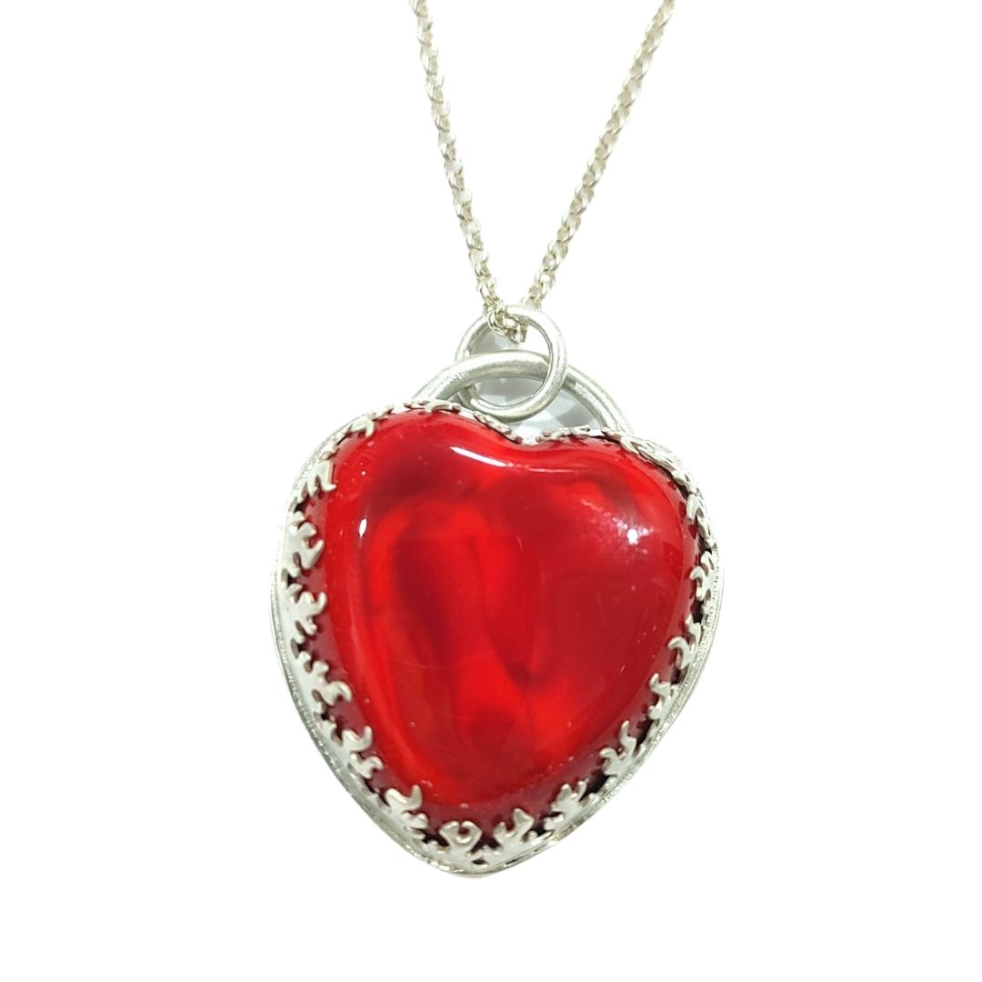 Pendant made from a large and thick lampwork glass stone in the shape of a heart. The heart is bright and bold red with swirls of lighter and darker reds, and is very shiny. The bezel that holds the glass stone is a cutout pattern of flames. All of the metal is sterling silver, and the pendant comes on a sterling silver necklace. Front view.