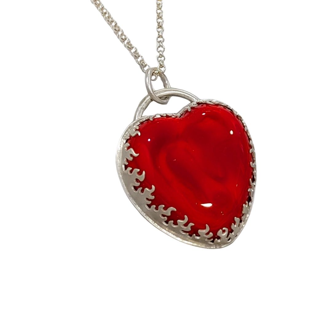Pendant made from a large and thick lampwork glass stone in the shape of a heart. The heart is bright and bold red with swirls of lighter and darker reds, and is very shiny. The bezel that holds the glass stone is a cutout pattern of flames. All of the metal is sterling silver, and the pendant comes on a sterling silver necklace. Front angled view showing some of the flame detail.