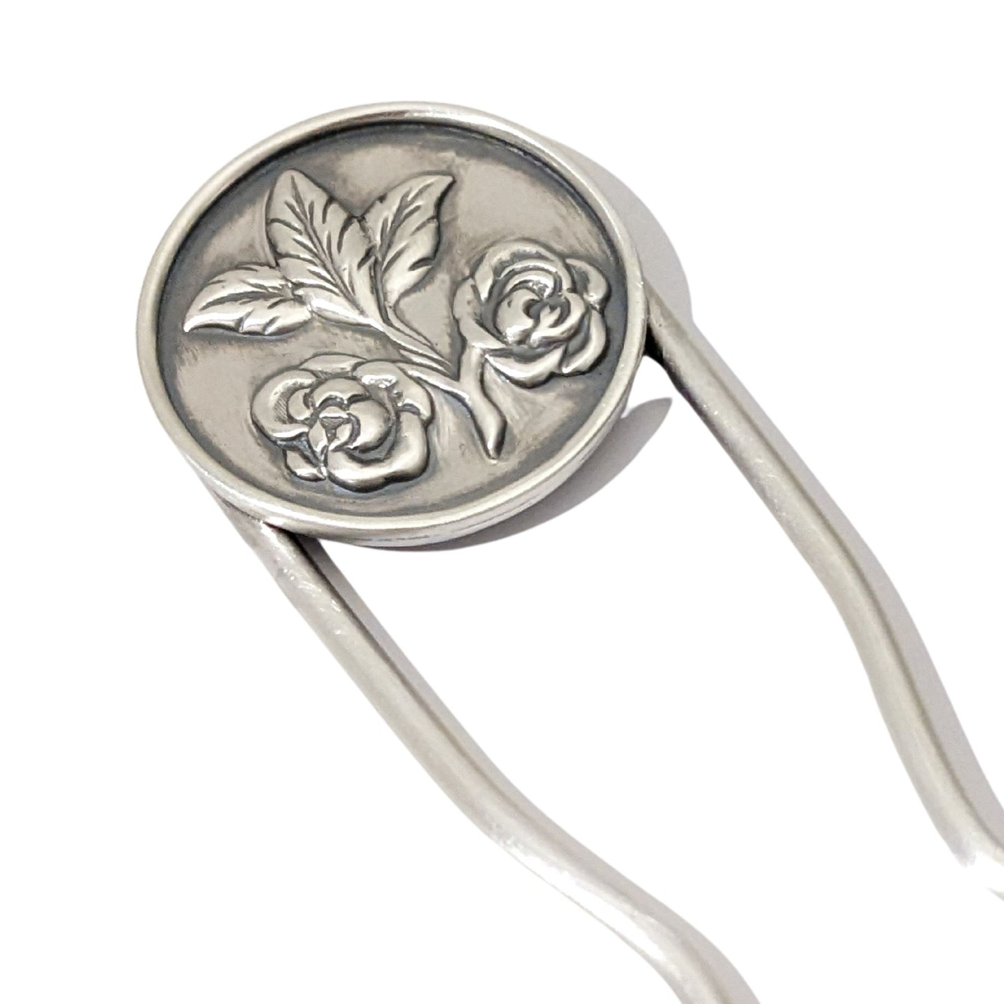 Sterling silver hair fork. Attached to top of fork is a round piece of silver with a rounded border edge. The sheet has a raised design of two roses growing off of a branch clipping and at the end of the branch there are three leaves.