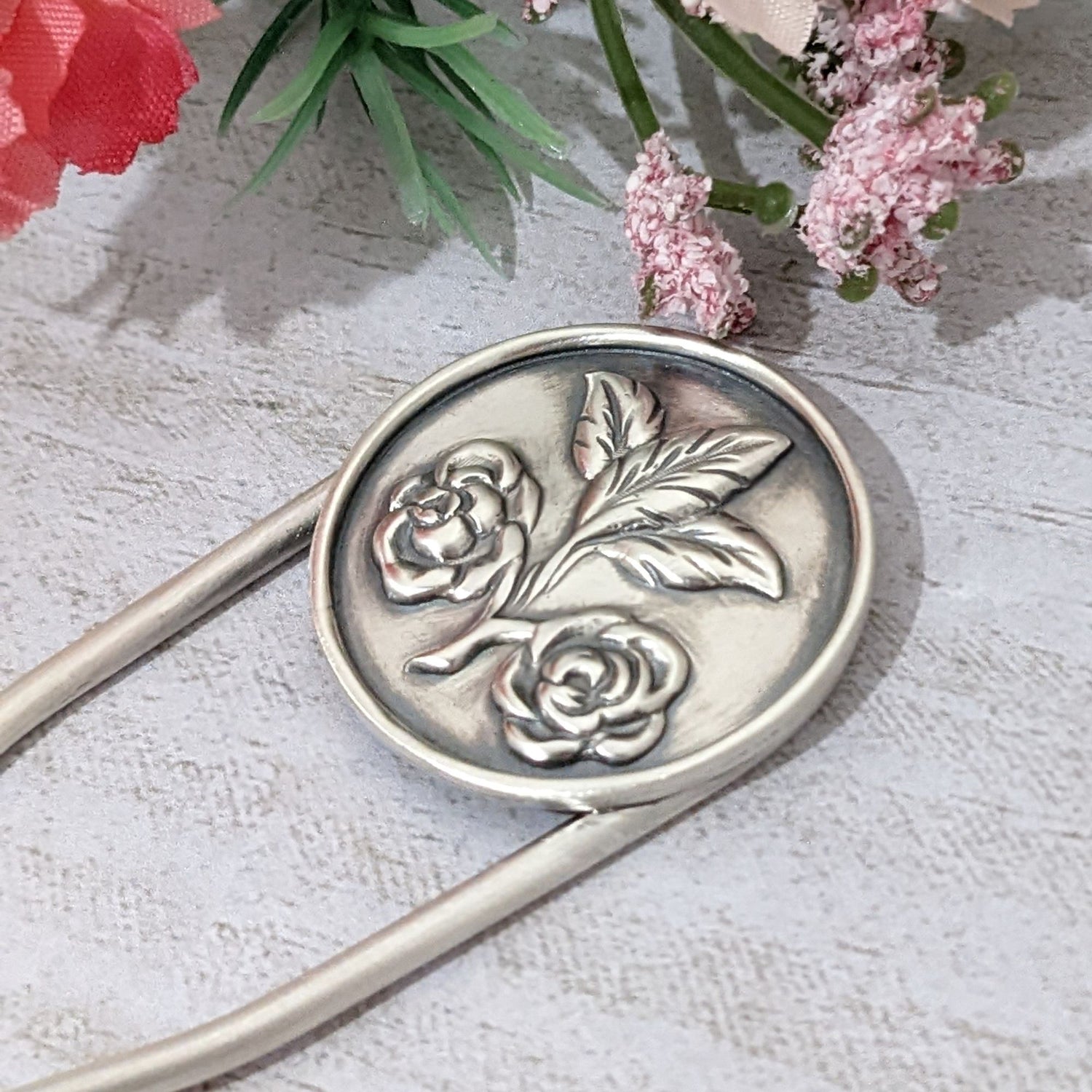 Sterling silver hair fork. Attached to top of fork is a round piece of silver with a rounded border edge. The sheet has a raised design of two roses growing off of a branch clipping and at the end of the branch there are three leaves. Staged photo on a gray background with flowers.