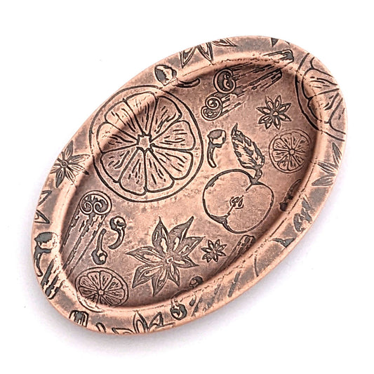 Copper oval ring dish with design of ingredients used in winter spice drinks, including apples oranges nutmeg cinnamon and clove. Dish is 2 inches by three inches with a raised lip.