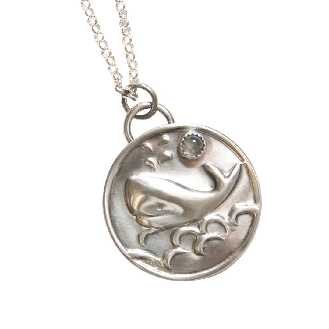 Round sterling silver pendant with a raised impression of a smiling whale. There are waves below the whale and a water spout above the whale. A labradorite gemstone is set in a cabochon style just to the right of the water spout. A half round loop above the disk holds a jump ring used for the sterling silver necklace that comes with the pendant.