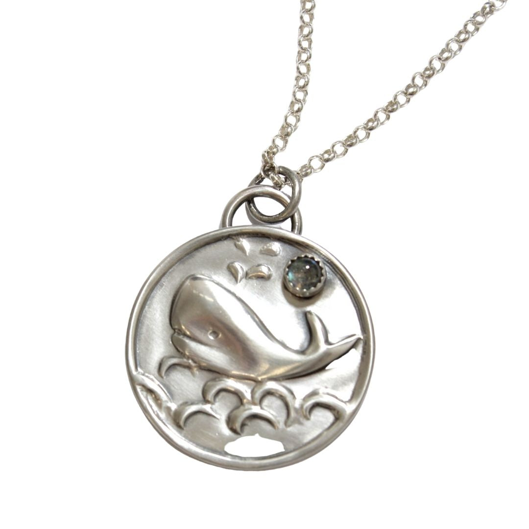 Round sterling silver pendant with a raised impression of a smiling whale. There are waves below the whale and a water spout above the whale. A labradorite gemstone is set in a cabochon style just to the right of the water spout. A half round loop above the disk holds a jump ring used for the sterling silver necklace that comes with the pendant.