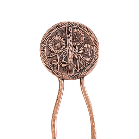Copper hair fork with a large round design at top of sunflowers and a peace sign. The peace sign has a wood texture. Three sunflowers grow through the wood sign. There's a lot of detail - veins on the leaves, you can see the sunflower seeds and petals. On a copper fork with a hammered finish.