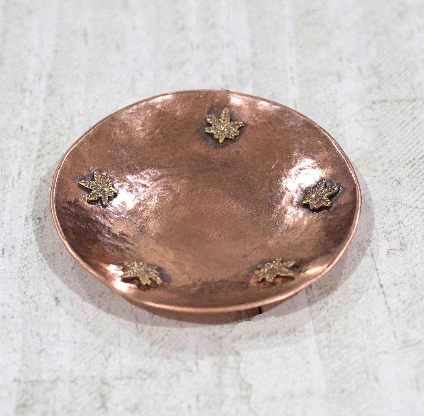 Cannabis Leaf Copper Bowl for rings, jewelry, trinkets. This small bowl has hammered texture and raised edges. Toward the edge of the bowl are five bronze cannabis leaves that are soldered to the copper.