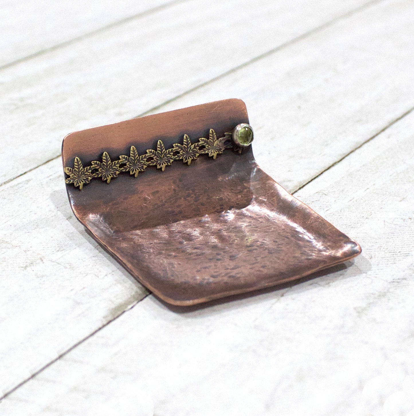 Small copper ring dish. Square shape, one edge is bent to form a short wall. On the wall is a row of 6 bronze cannibas leaves that are soldered to the copper. At the right edge of the leaves there is a small silver setting with a green peridot gemstone.