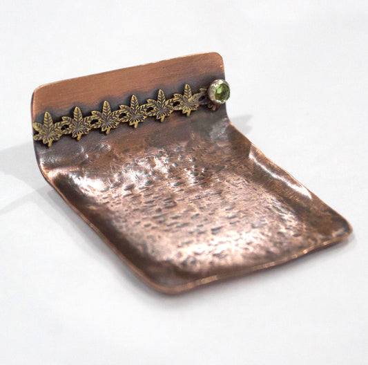 Small copper ring dish. Square shape, one edge is bent to form a short wall. On the wall is a row of 6 bronze cannibas leaves that are soldered to the copper. At the right edge of the leaves there is a small silver setting with a green peridot gemstone.