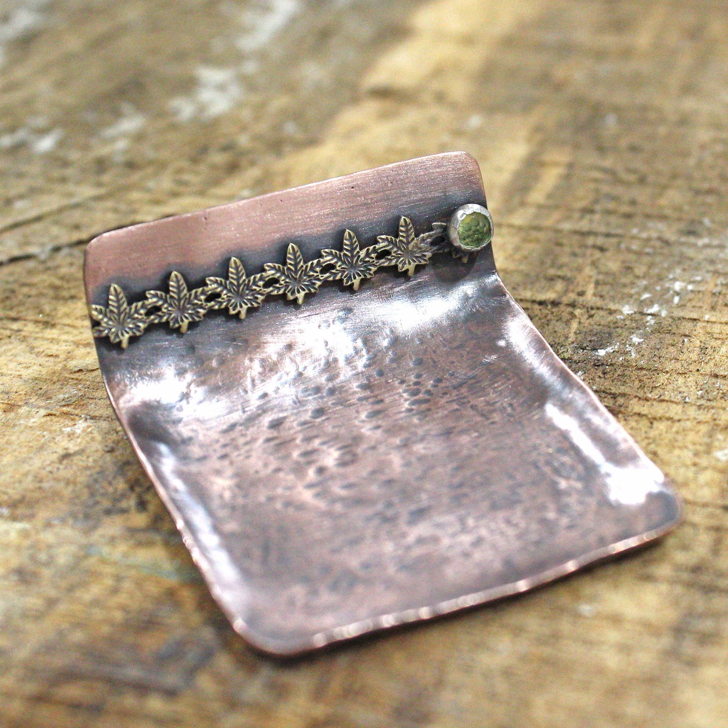 Small copper ring dish. Square shape, one edge is bent to form a short wall. On the wall is a row of 6 bronze cannibas leaves that are soldered to the copper. At the right edge of the leaves there is a small silver setting with a green peridot gemstone.