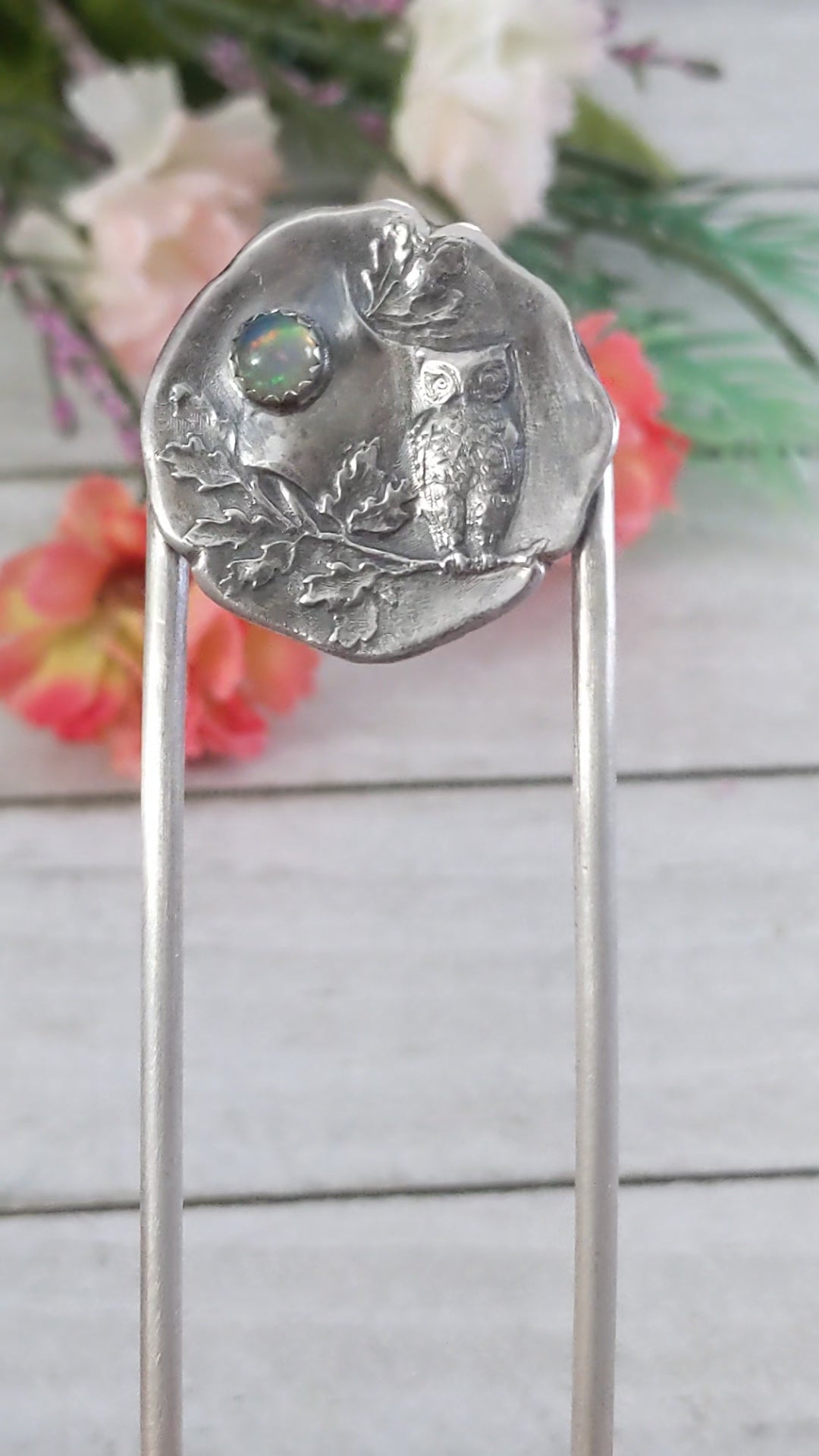 Video of Sterling silver hair fork. The design at the top of the hair fork is a round sterling silver piece with a 3 dimensional design of an owl sitting on a tree branch. The branch has leaves, oak perhaps. Above the leaves there is a white opal gemstone to represent the moon. 