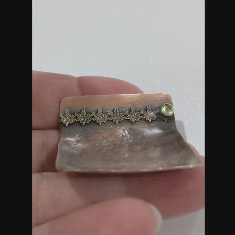 Small copper ring dish. Square shape, one edge is bent to form a short wall. On the wall is a row of 6 bronze cannibas leaves that are soldered to the copper. At the right edge of the leaves there is a small silver setting with a green peridot gemstone.