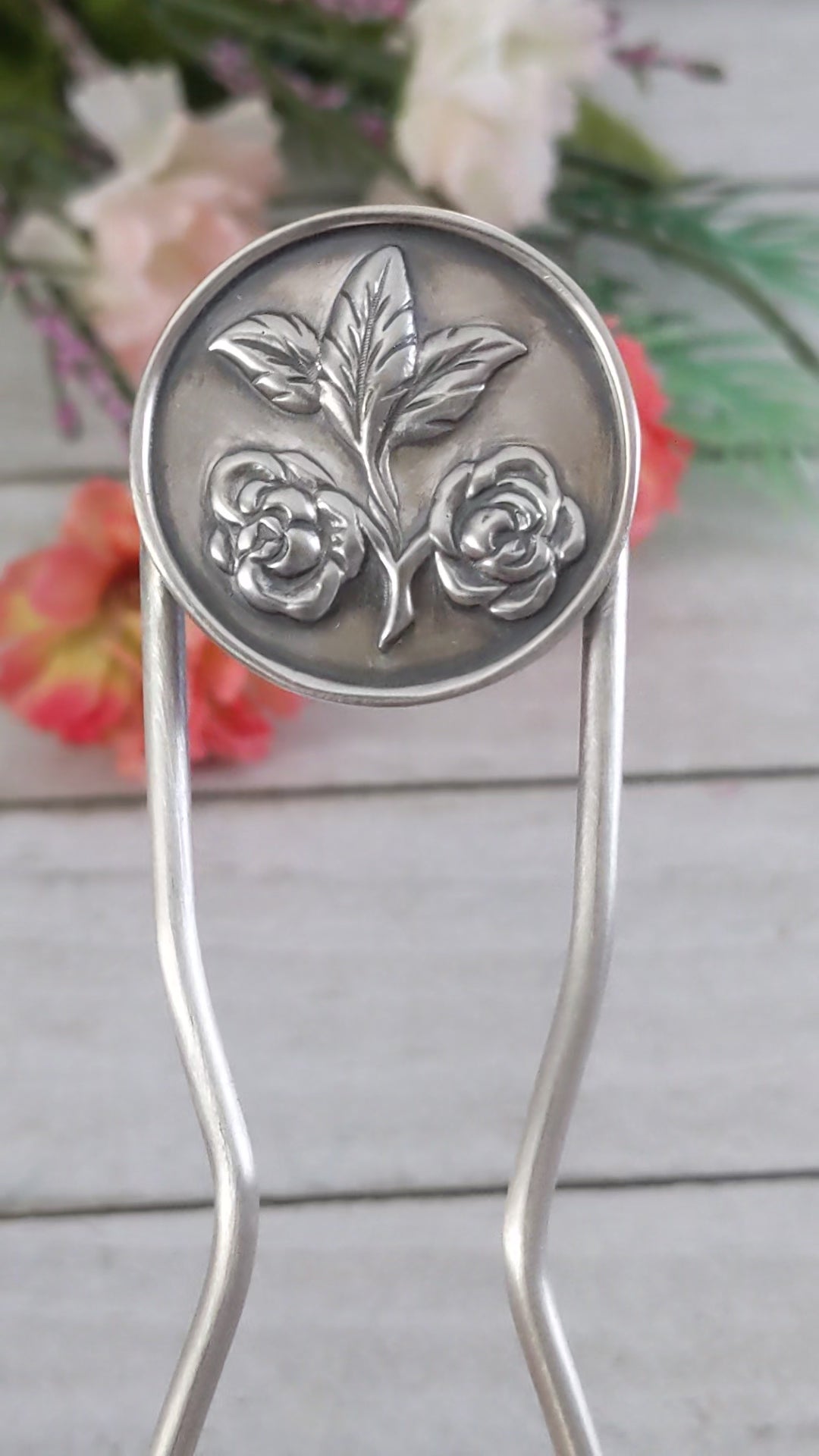 Sterling silver hair fork. Attached to top of fork is a round piece of silver with a rounded border edge. The sheet has a raised design of two roses growing off of a branch clipping and at the end of the branch there are three leaves.