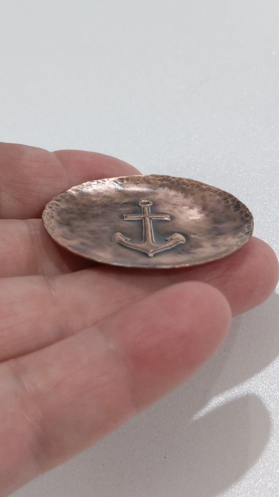 Copper ring or jewelry or trinket dish with a raised impression of a boat anchor. The edged of the dish are curved up slightly, the copper metal has a hammered texture, and the dish is oxidized to enhance the detail.