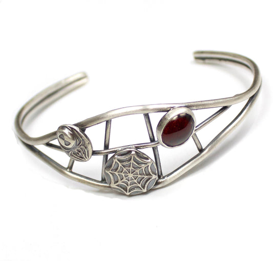 Sterling silver cuff bracelet. There are two sturdy silver wires that separate in the middle part of the cuff. Between the wires there are smaller silver wires in a spider web design. On top of the web design there is a silver spider, a smaller silver web, and an oval red garnet cabochon gemstone. 