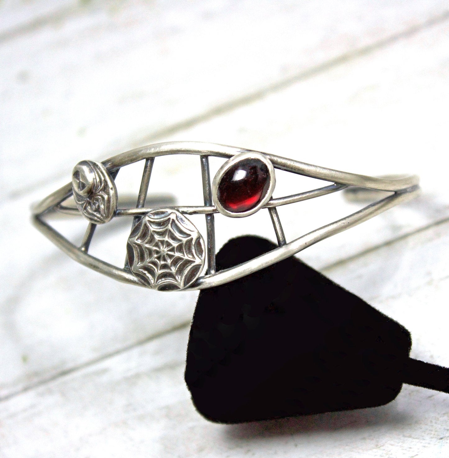 Sterling silver cuff bracelet. There are two sturdy silver wires that separate in the middle part of the cuff. Between the wires there are smaller silver wires in a spider web design. On top of the web design there is a silver spider, a smaller silver web, and an oval red garnet cabochon gemstone. 