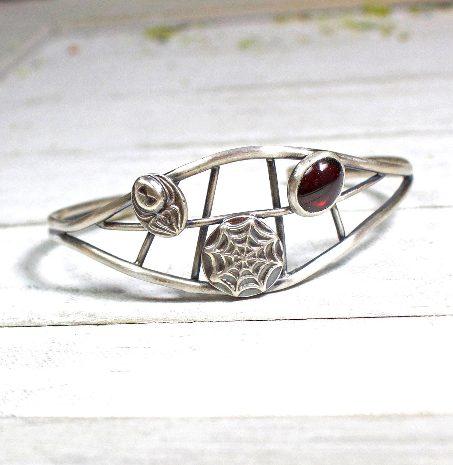 Sterling silver cuff bracelet. There are two sturdy silver wires that separate in the middle part of the cuff. Between the wires there are smaller silver wires in a spider web design. On top of the web design there is a silver spider, a smaller silver web, and an oval red garnet cabochon gemstone. 