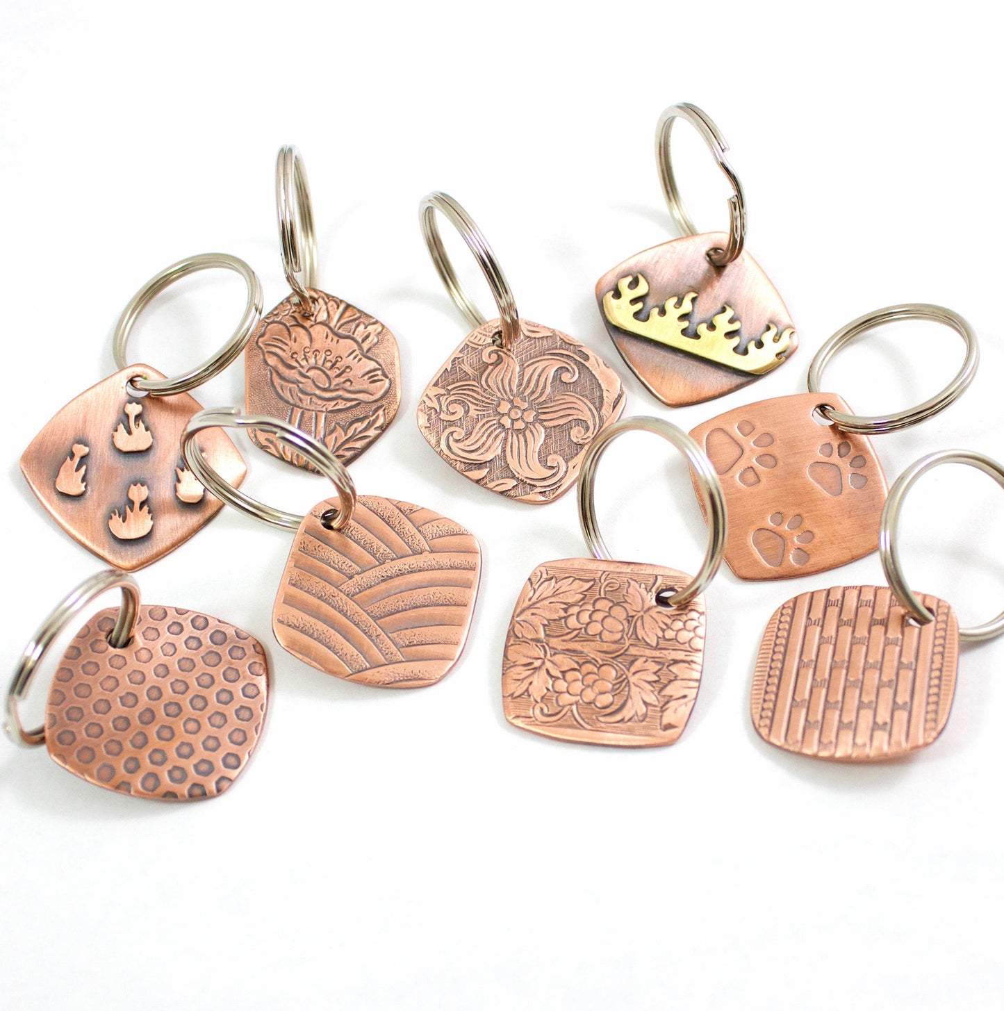 Collection of copper keychains, all square with softly rounded corners. Desgins include flowers, flames, pawprints, bricks, grapes, and arches