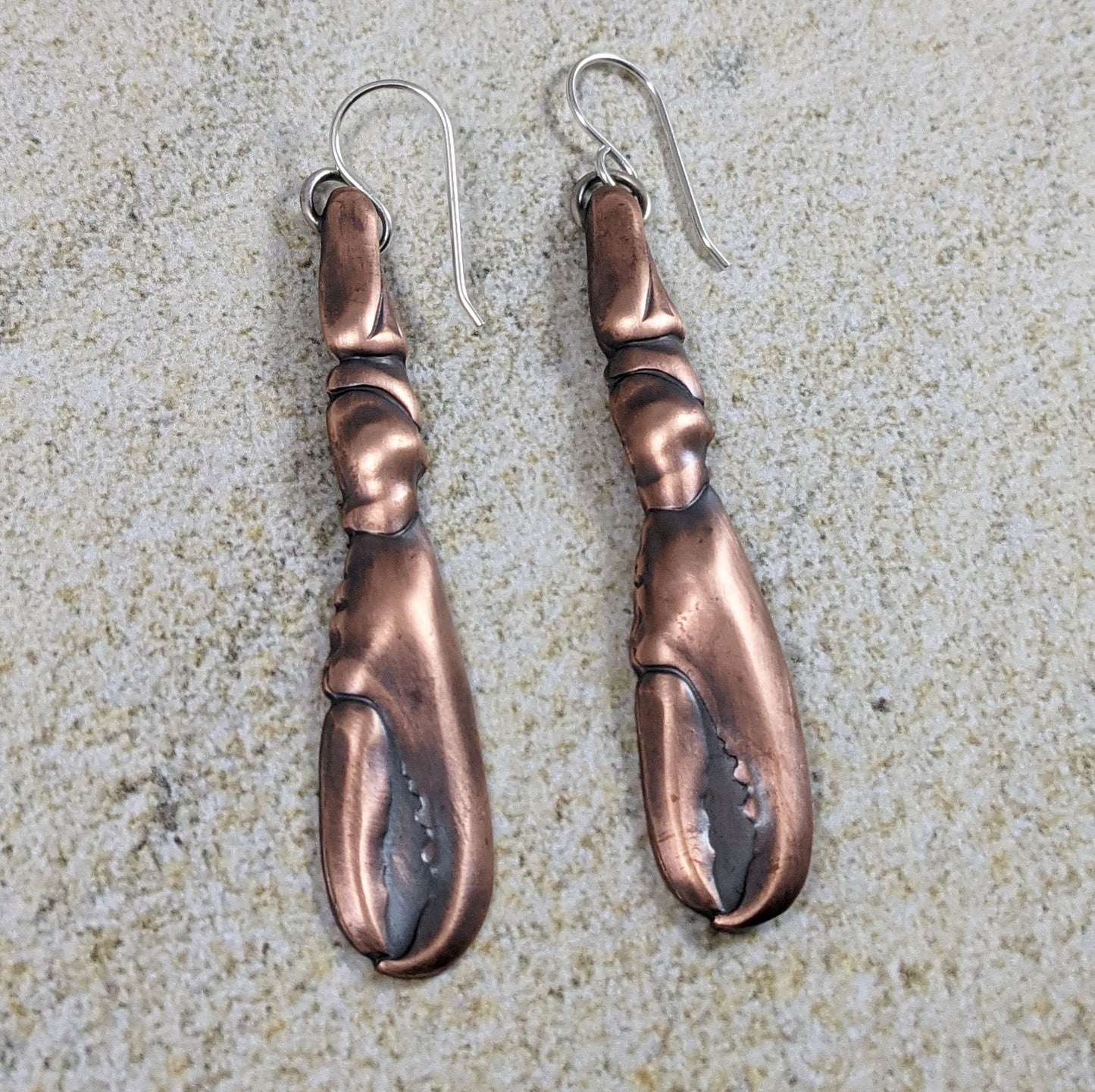 Copper earrings in the shape of a lobster claw. The claws are three dimensional and are on sterling silver ear wires. Posed on a sandy background.