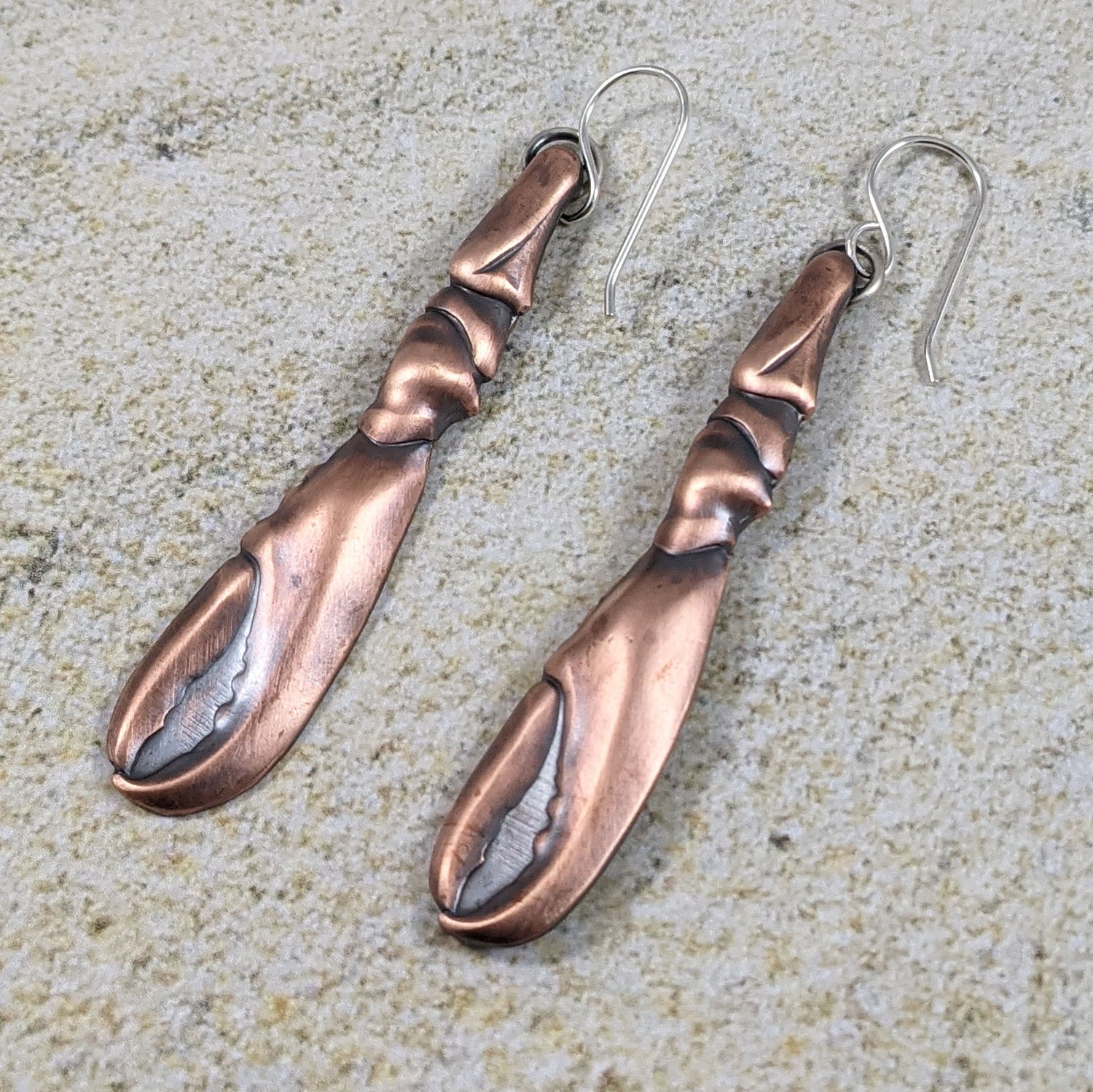 Copper earrings in the shape of a lobster claw. The claws are three dimensional and are on sterling silver ear wires. Posed on a sandy background.